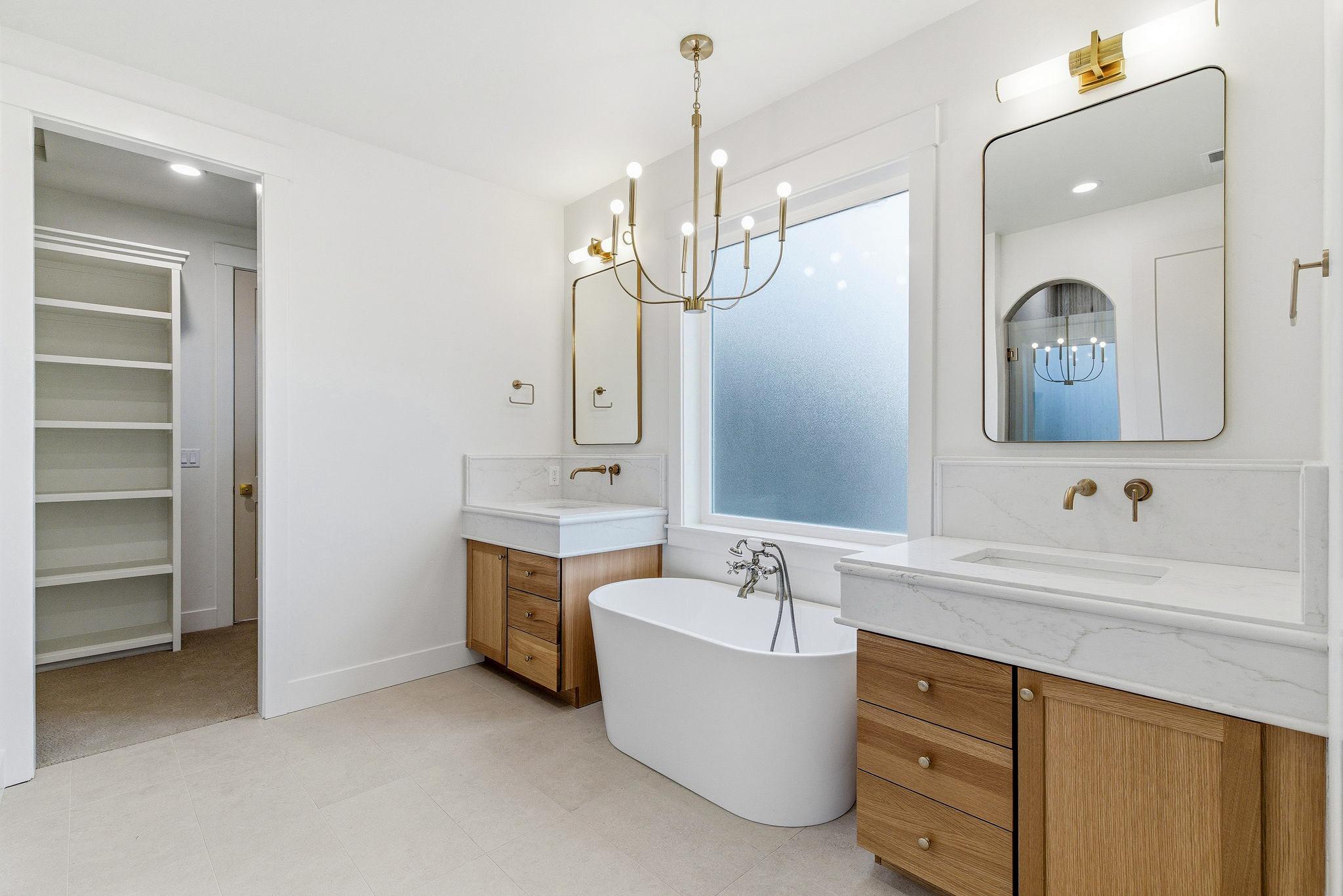 The primary bath: freestanding tub, dual white oak vanities, brass fixtures, and a walk-in closet beyond