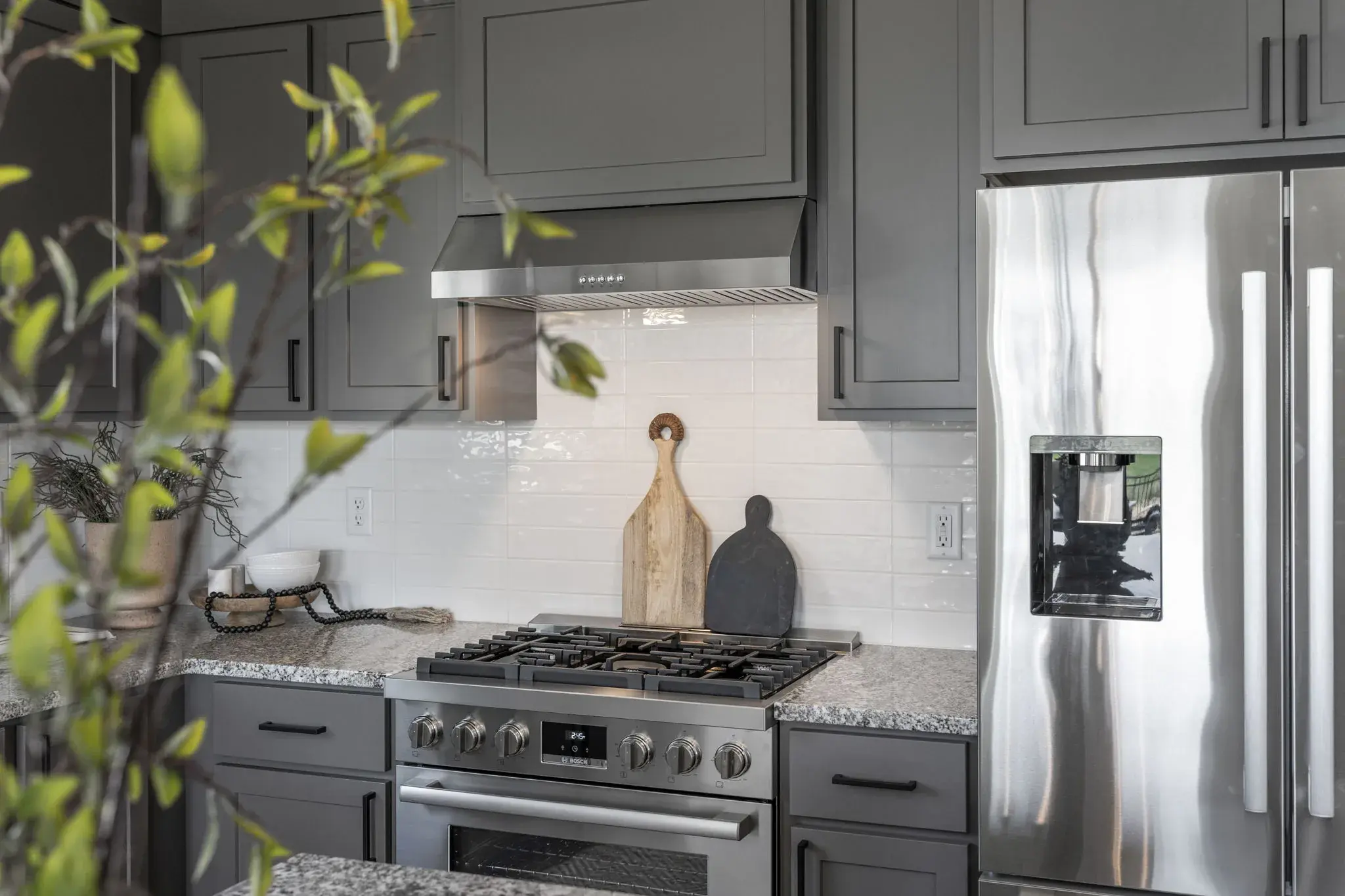 The gas range and tiled backsplash in the kitchen at Harvest Creek Vaulted Home, between grey cabinetry and a stainless refrigerator