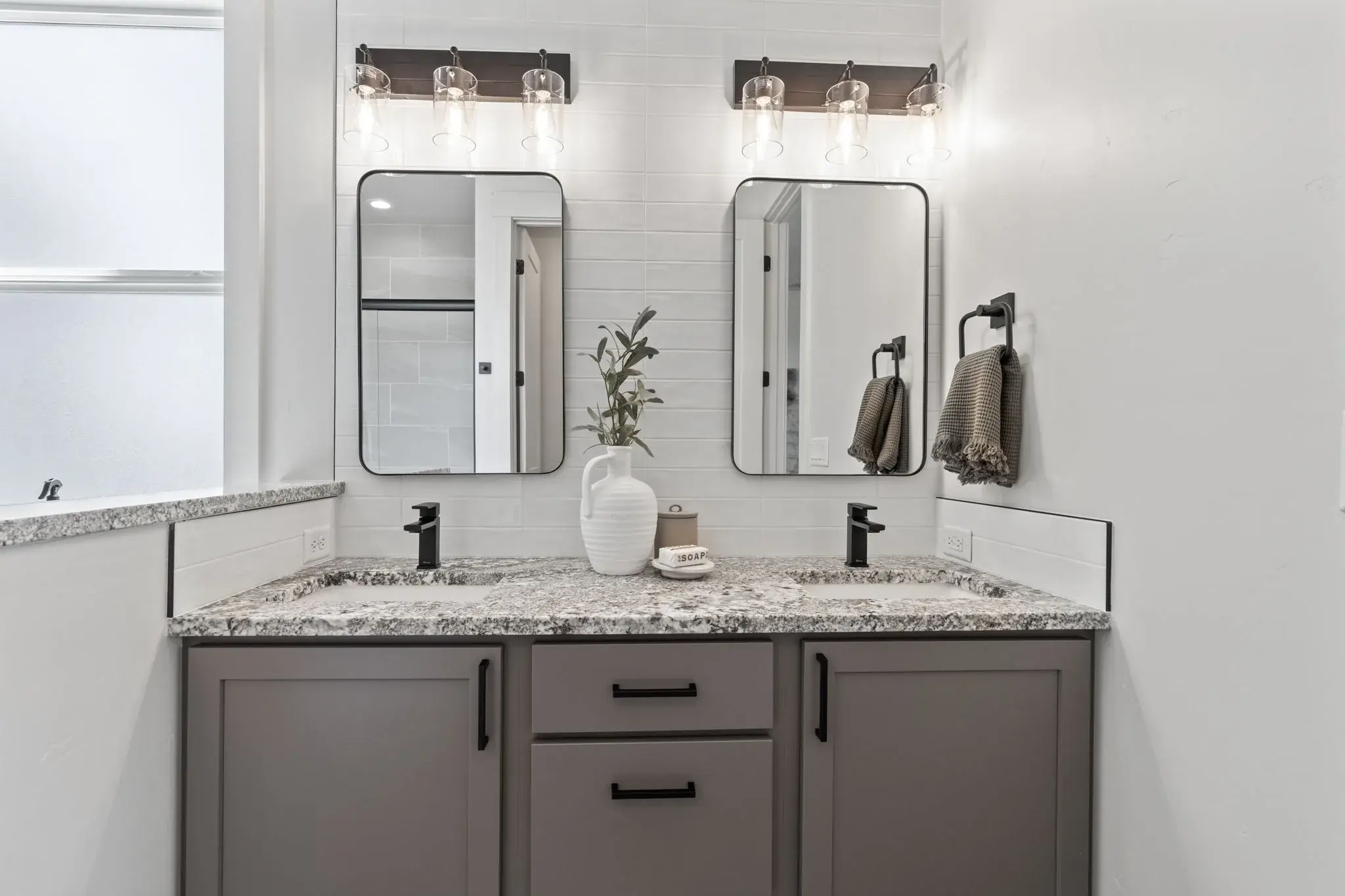 The double granite vanity in the primary bathroom at Harvest Creek Vaulted Home, with twin mirrors and glass sconces